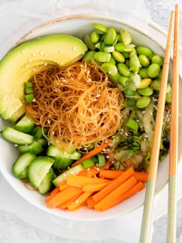 Close up of a white bowl with rice noodles surrounded by edamame, sliced carrots, cucumbers, and avocado topped with sesame seeds