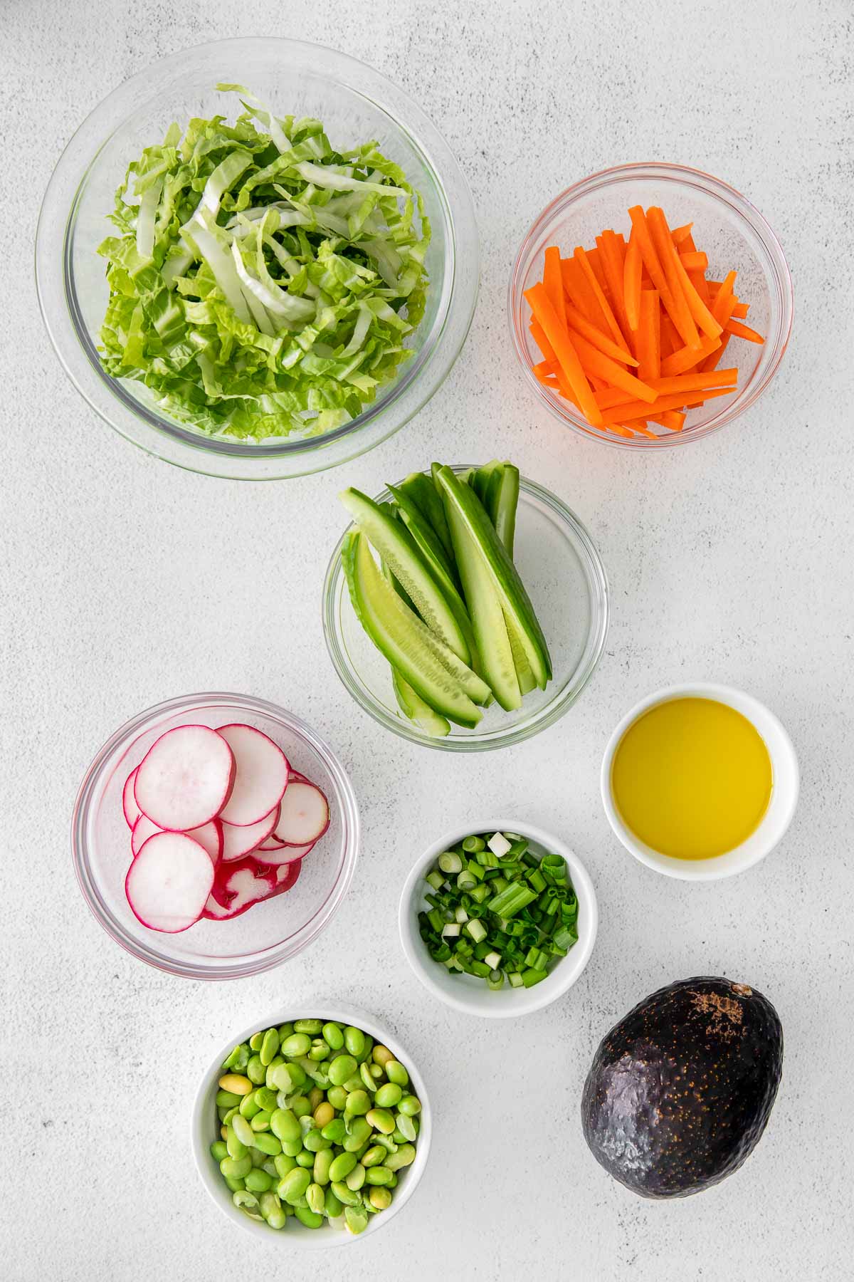 several small glass bowls of edamame, sliced radishes, sliced green onions, cabbage, sliced carrots and a whole avocado.