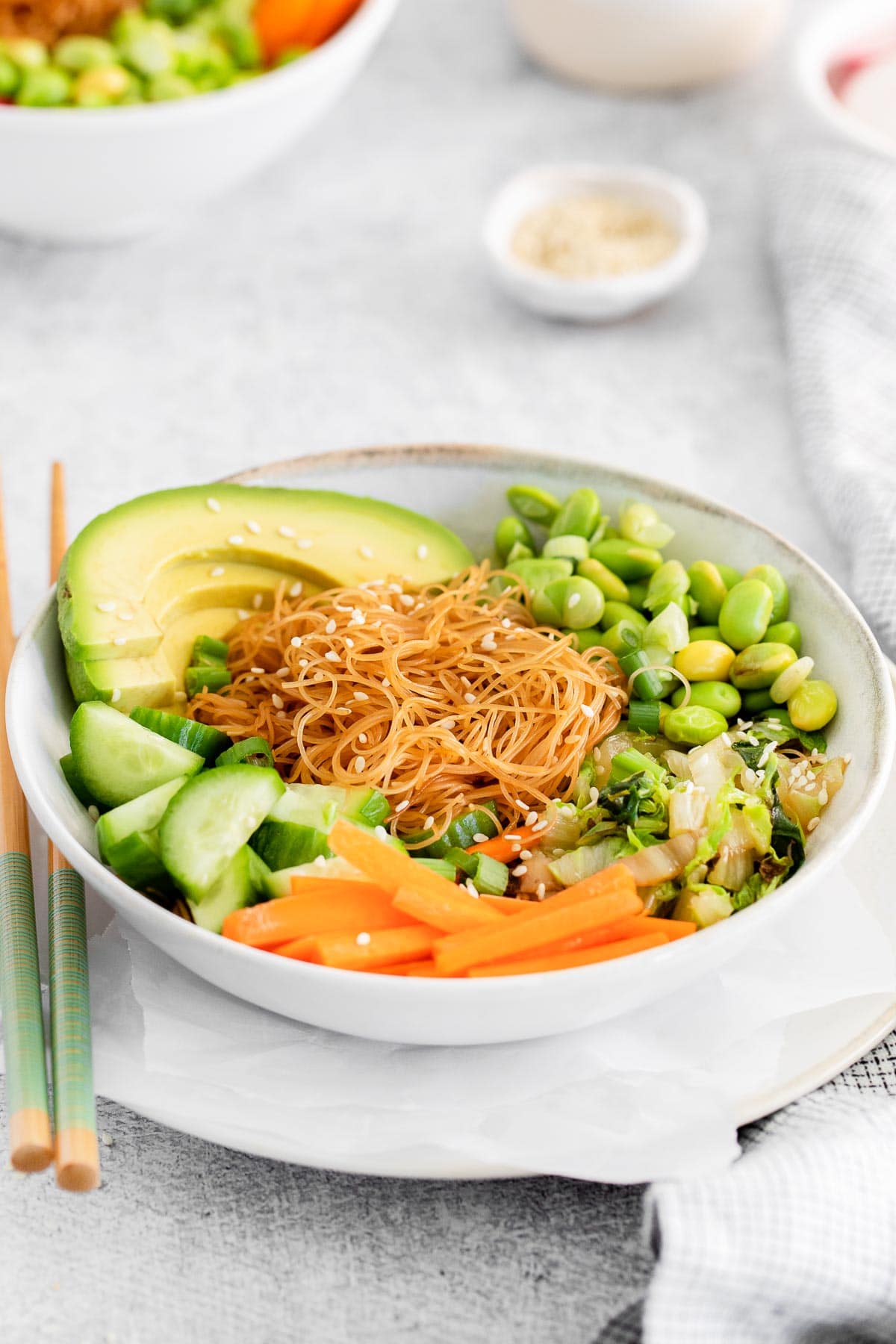white bowl with sliced carrots, edamame sliced cucumbers, rice noodles and avocado topped with sesame seeds