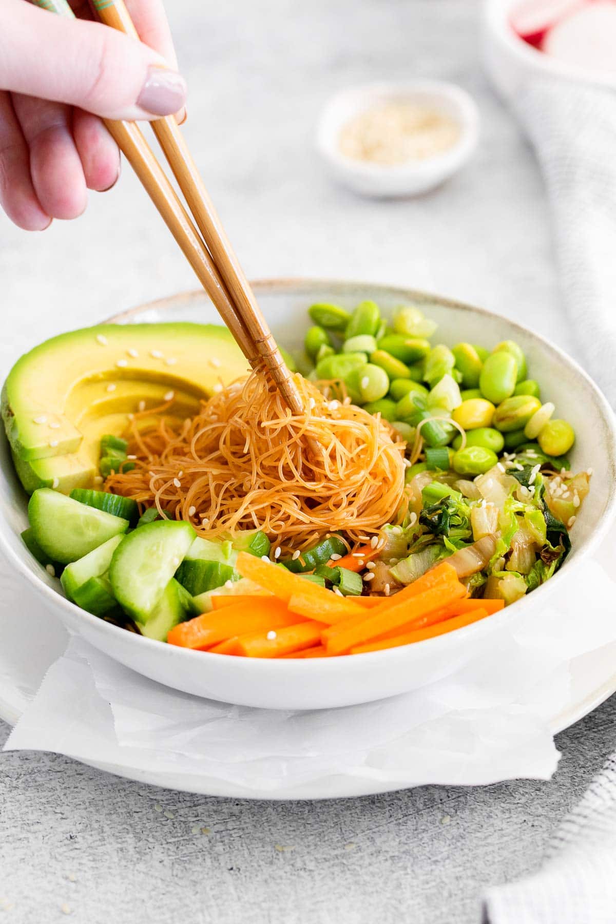 chopsticks picking up rice noodles from a white bowl full of sliced carrots, cucumbers, cabbage and avocado