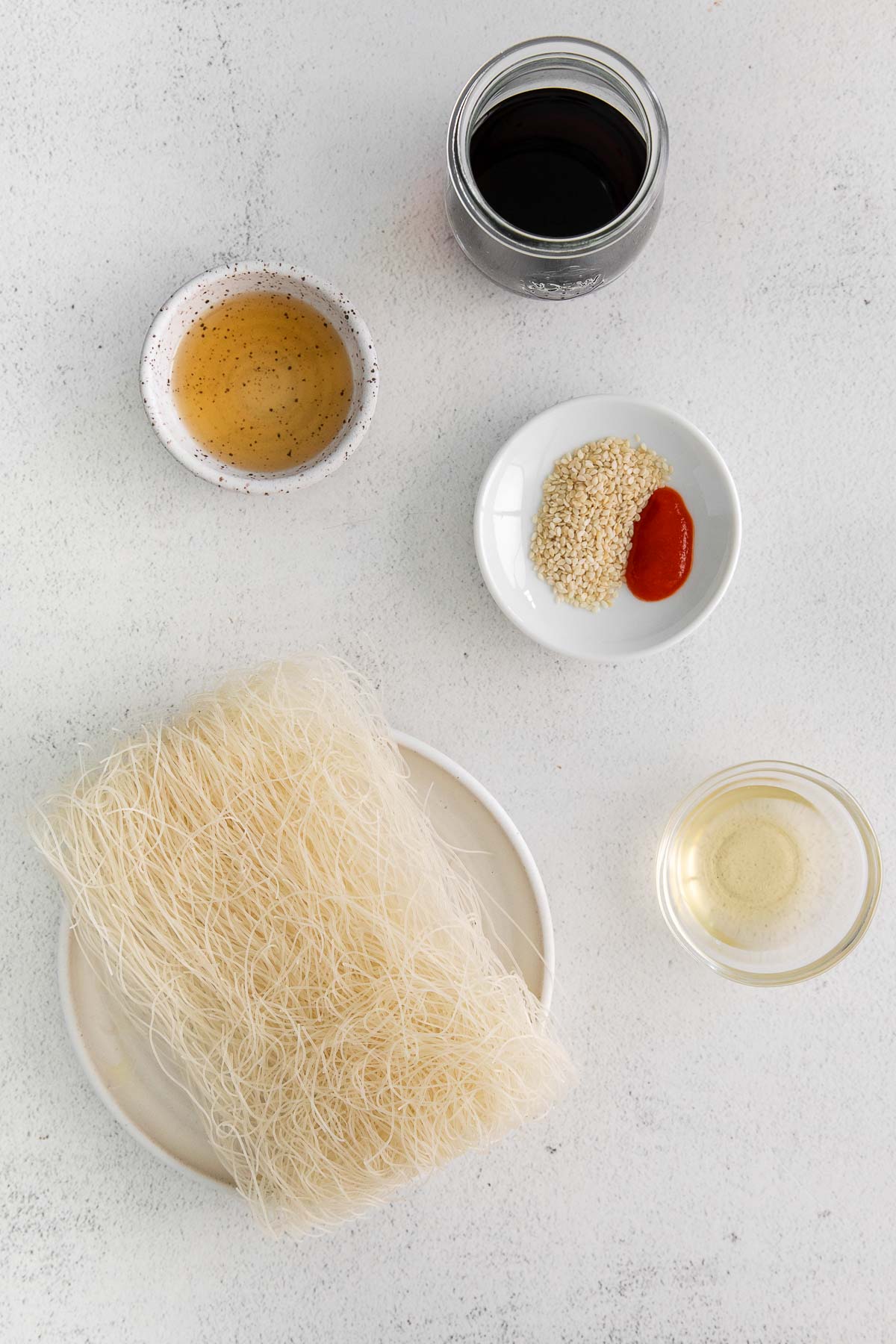 uncooked rice noodles on a white plate and small bowls of soy sauce, sesame oil and spices
