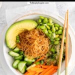 white bowl of edamame, rice noodles, avocado slices, carrots and cucumbers with chopsticks on the side