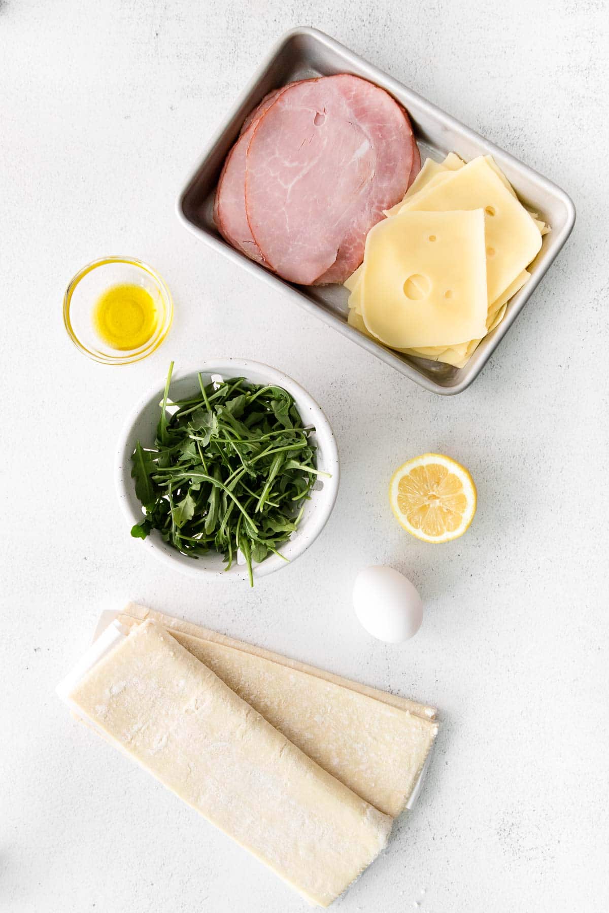 small silver tray with a stack of sliced ham and sliced cheese, puff pastry dough, a bowl of arugula, an egg, and olive oil