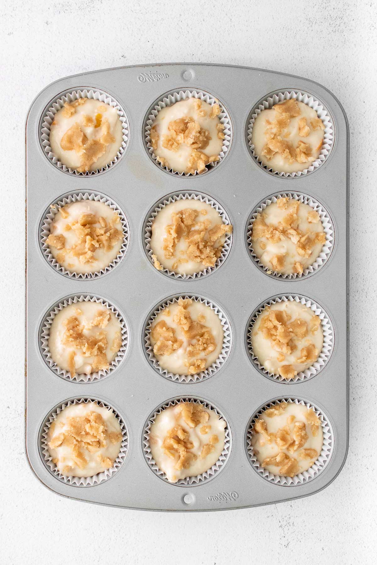 muffin tin filled with white paper liners with peach muffin batter topped with streusel topping