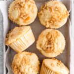 six peach muffins with streusel topping on a white parchment paper lined baking sheet