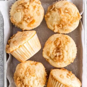 six peach muffins with streusel topping on a white parchment paper lined baking sheet