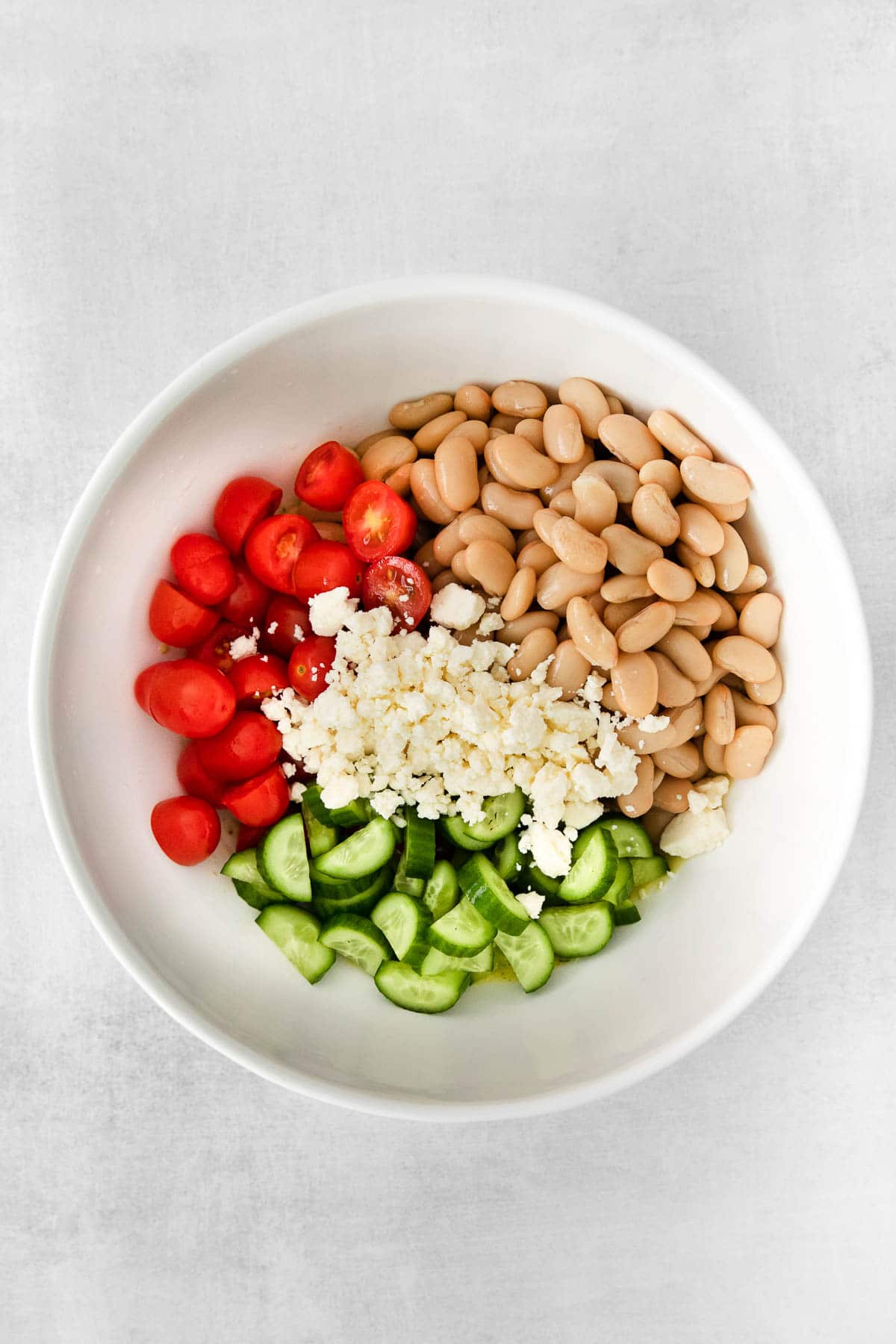 white bowl with white beans, diced cucumbers, sliced cherry tomatoes and feta cheese in four sections