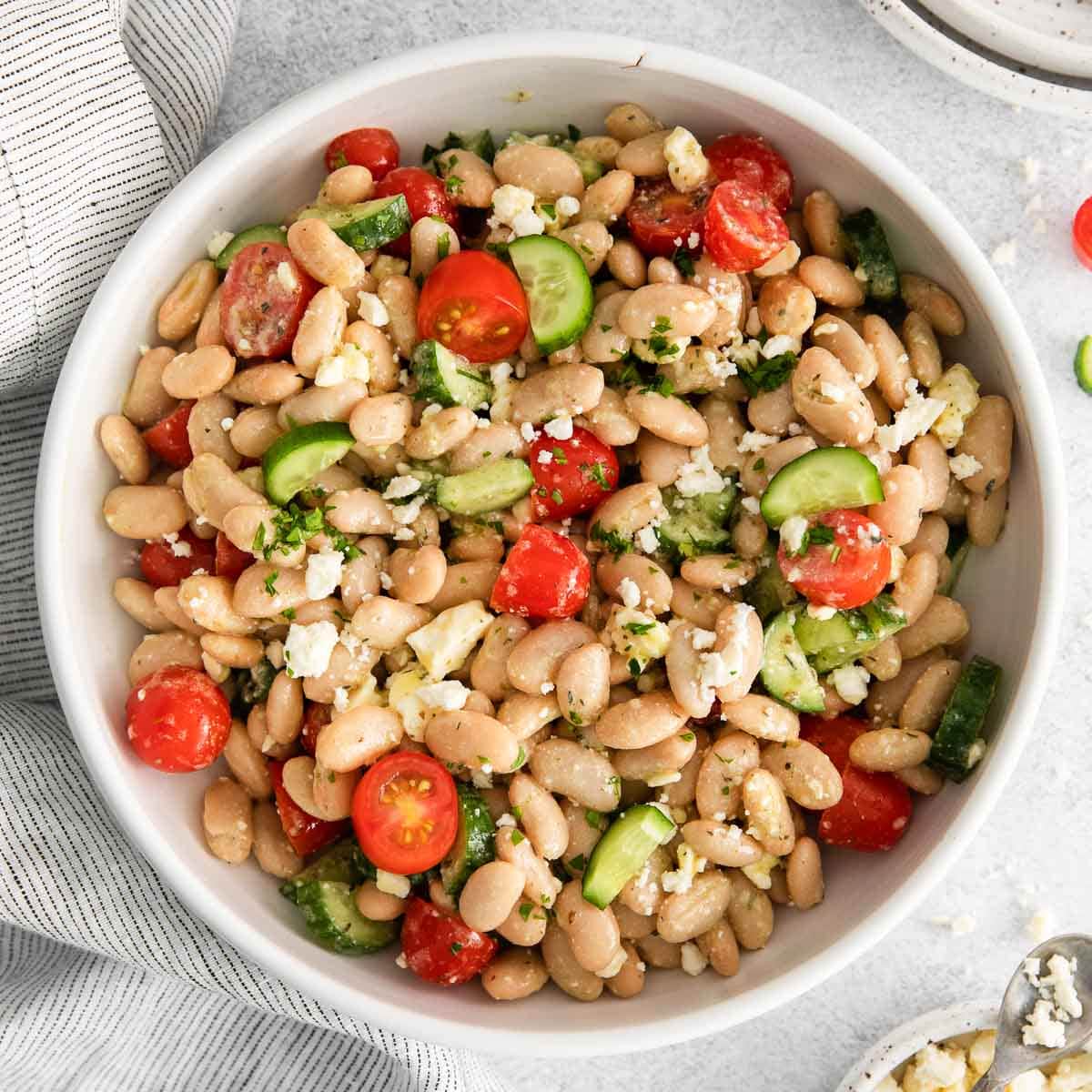 white bean salad with cherry tomatoes, cucumbers, feta cheese and greek spices in a white serving bowl