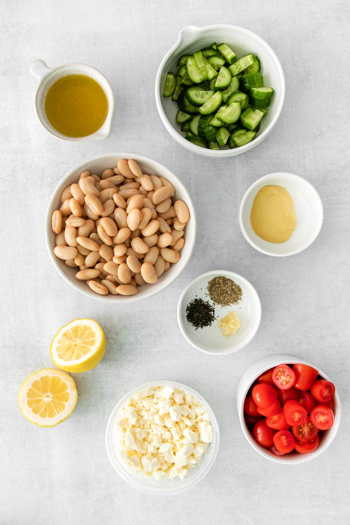 several white bowls with ingredients for white bean salad - cannellini beans, cherry tomatoes, diced cucumbers, feta cheese, lemon, dijon mustard, garlic and italian seasoning