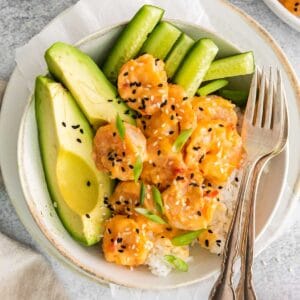 shrimp coated in bang bang sauce over rice with sliced avocado and sliced cucumber