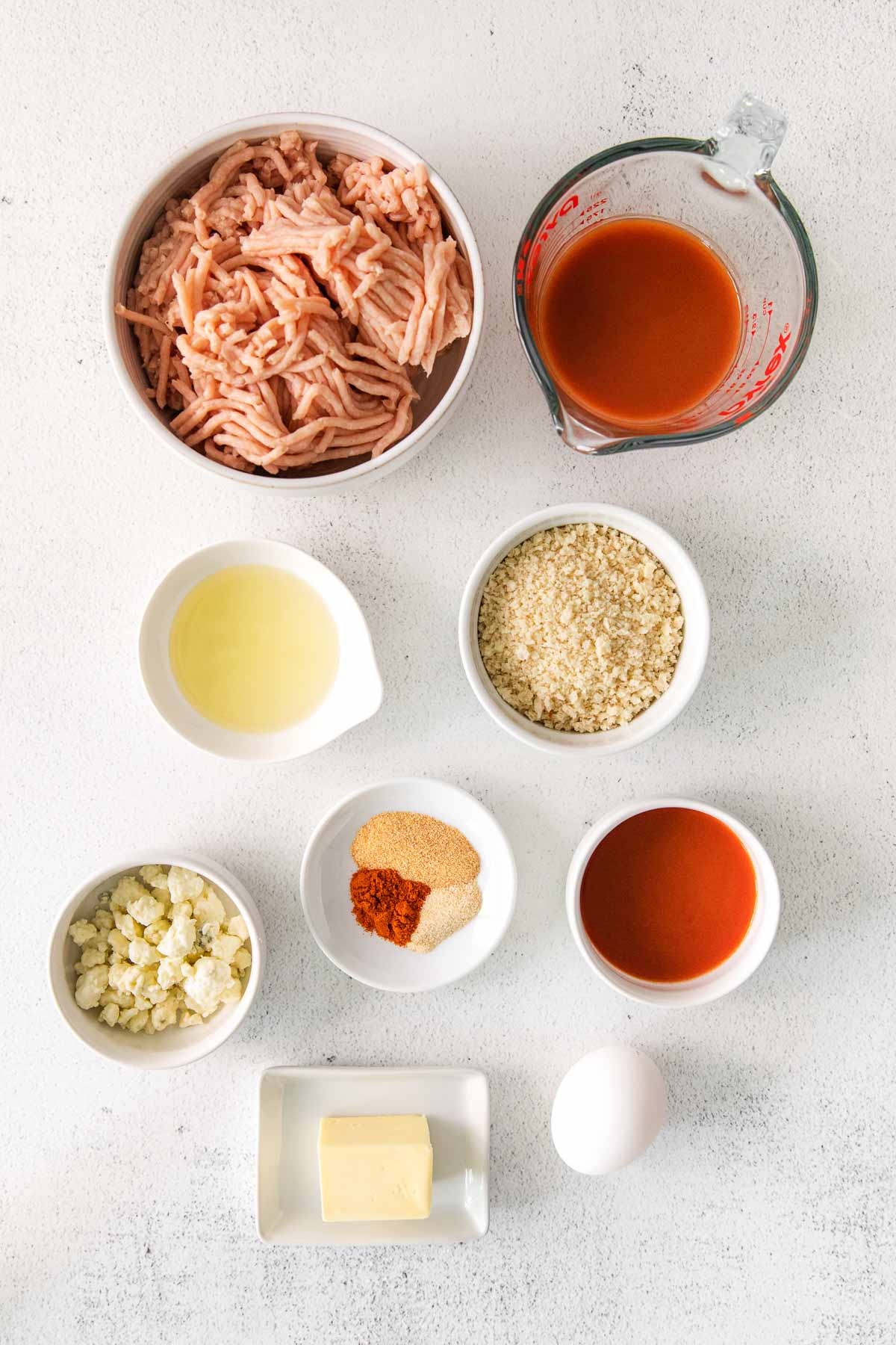 several white bowls with ingredient for buffalo chicken meatballs - raw ground chicken, buffalo sauce, butter, egg, pano breadcrumbs, blue cheese, spices and oils