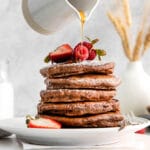 white plate with a stack of five chocolate pancakes topped with powdered sugar and strawberries and syrup being drizzled over top