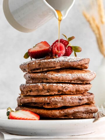 white plate with a stack of five chocolate pancakes topped with powdered sugar and strawberries and syrup being drizzled over top