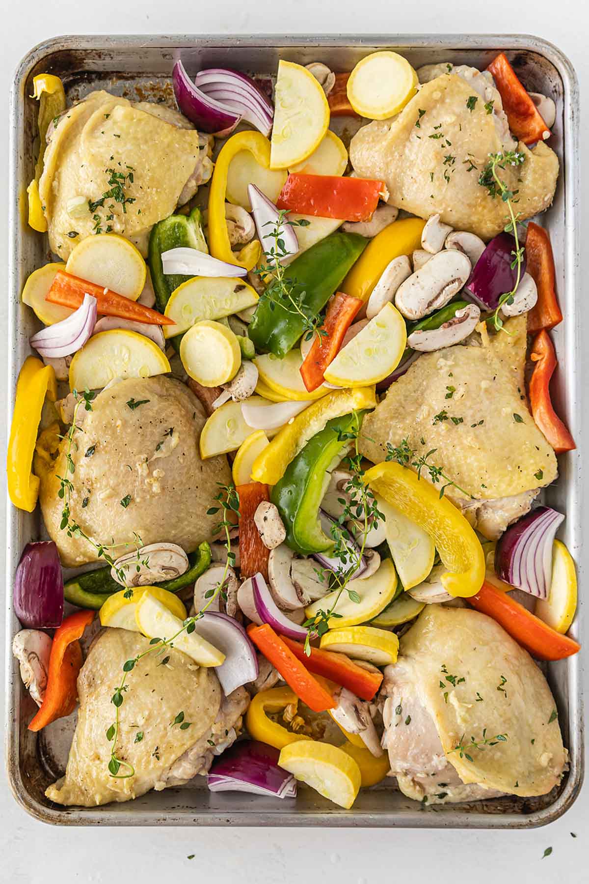 baking sheet with partly cooked chicken thighs surrounded by uncooked red bell peppers, yellow peppers, mushrooms, squash and red onions