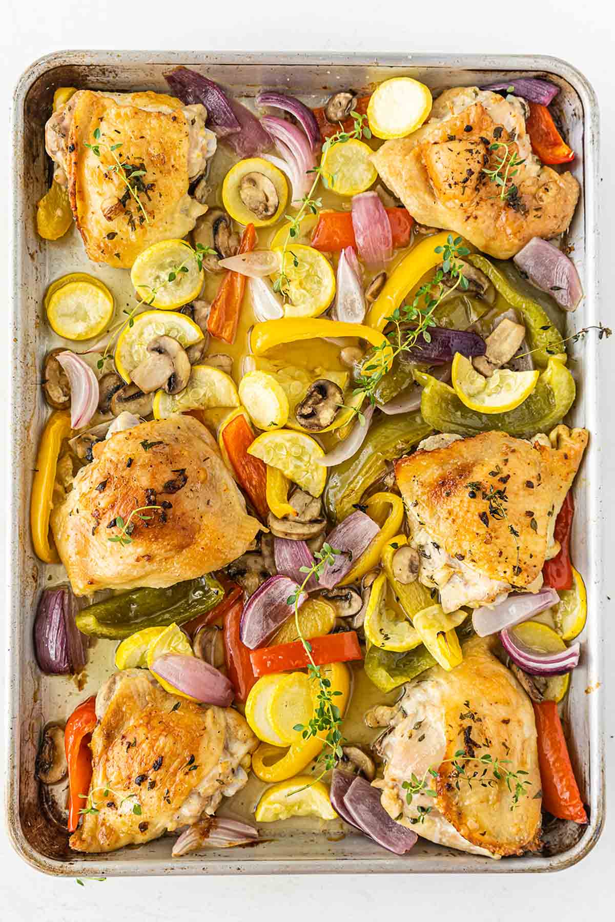 sheet pan full of oven baked chicken thighs and cooked red onion, red bell peppers, yellow squash and mushrooms.