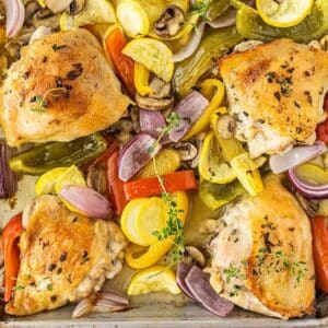 four crispy baked chicken thighs on a baking sheet with red bell peppers, mushroom and yellow squash.