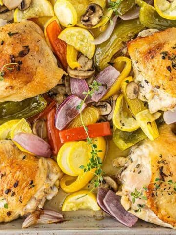 four crispy baked chicken thighs on a baking sheet with red bell peppers, mushroom and yellow squash.