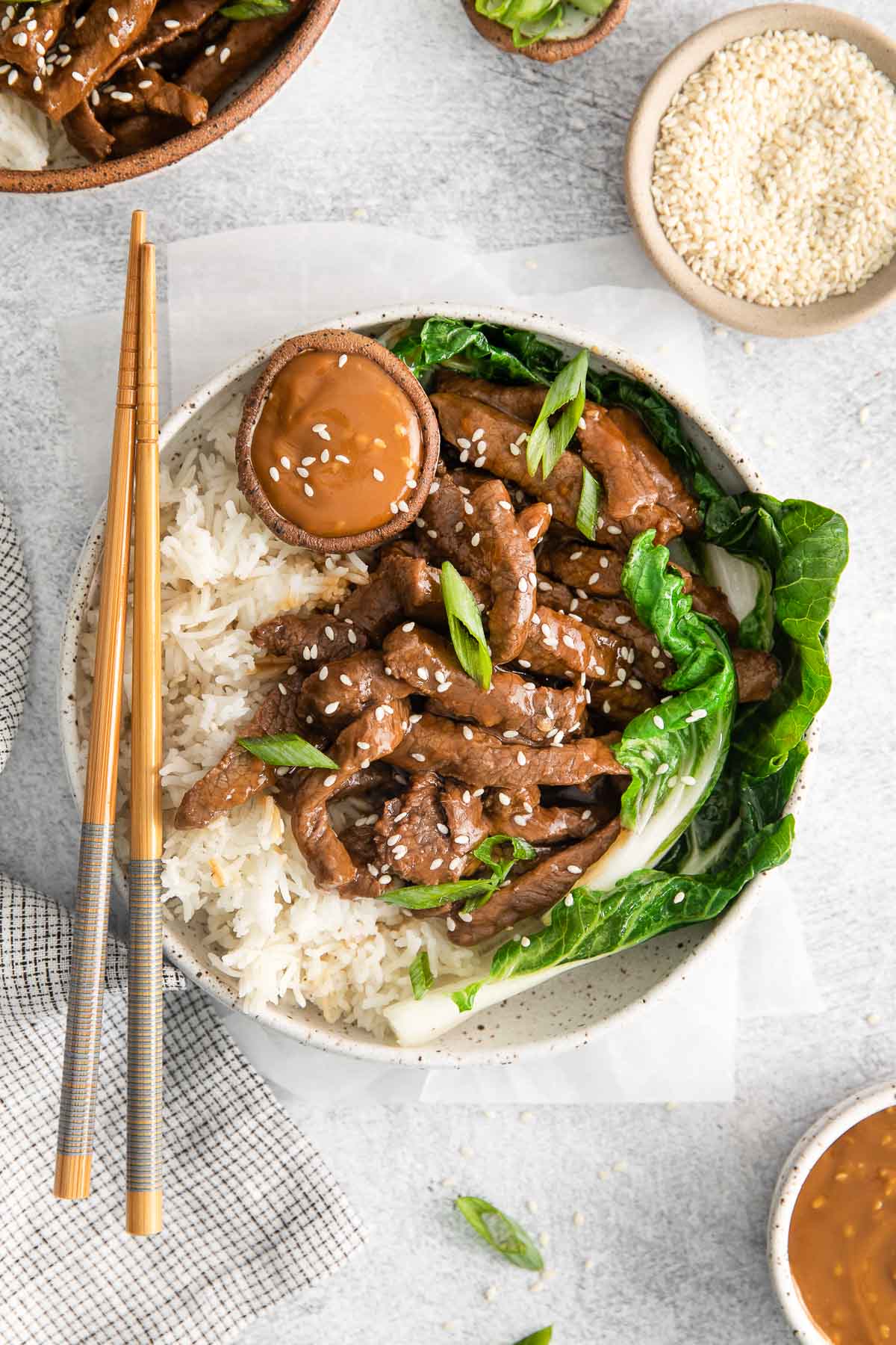 white bowl on a counter with white rice topped with strips of cooked steak with bok choy and sesame seeds with chop sticks on the side of the bowl.