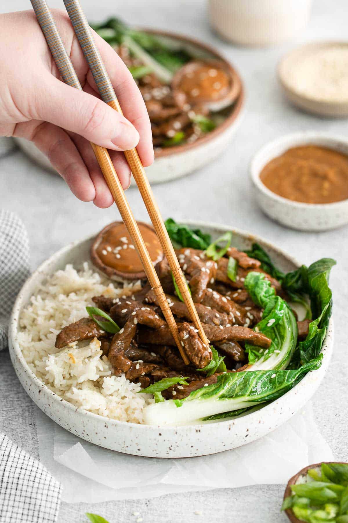 chop sticks picking up a strip of cooked steak in a bowl with white rice and bok choy.