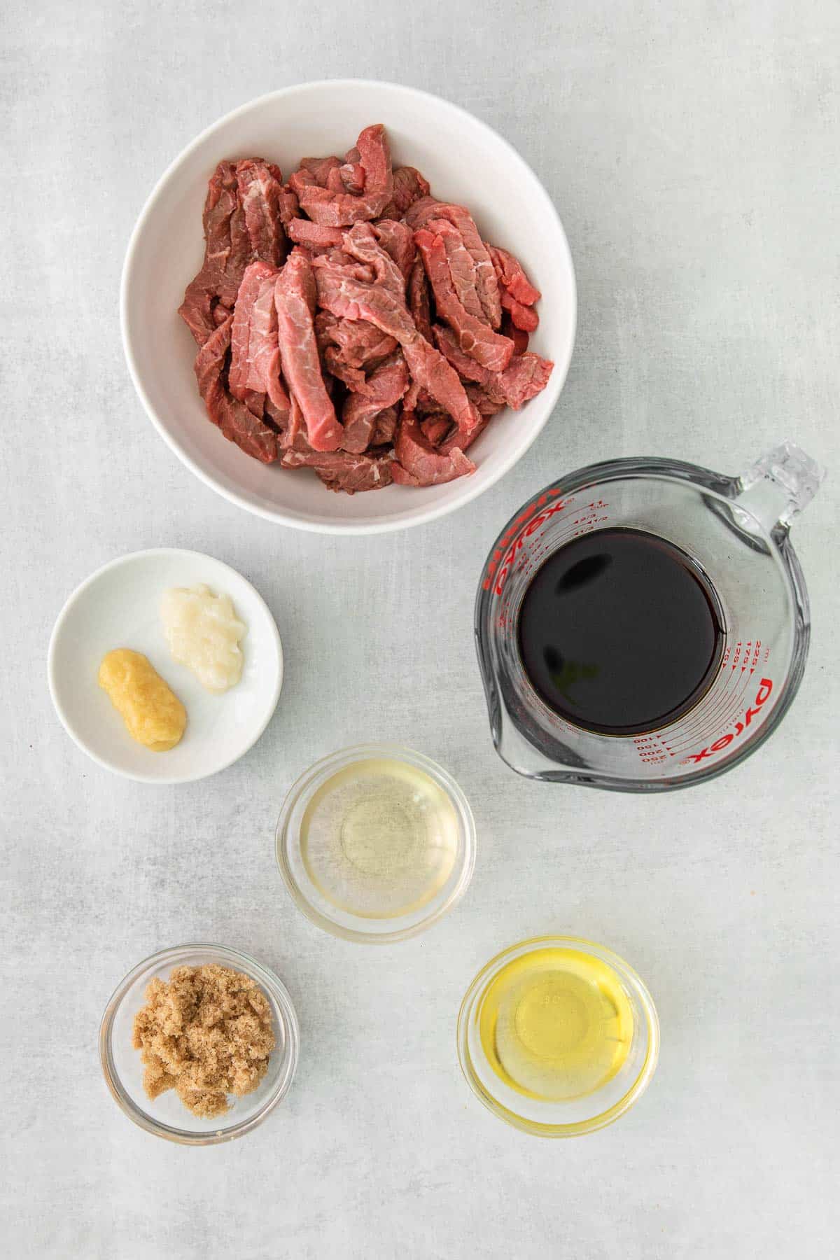 several glass bowls with ingredients for bulgogi beef - raw strips of steak, brown sugar, olive oil , rice vinegar, soy sauce, garlic and ginger