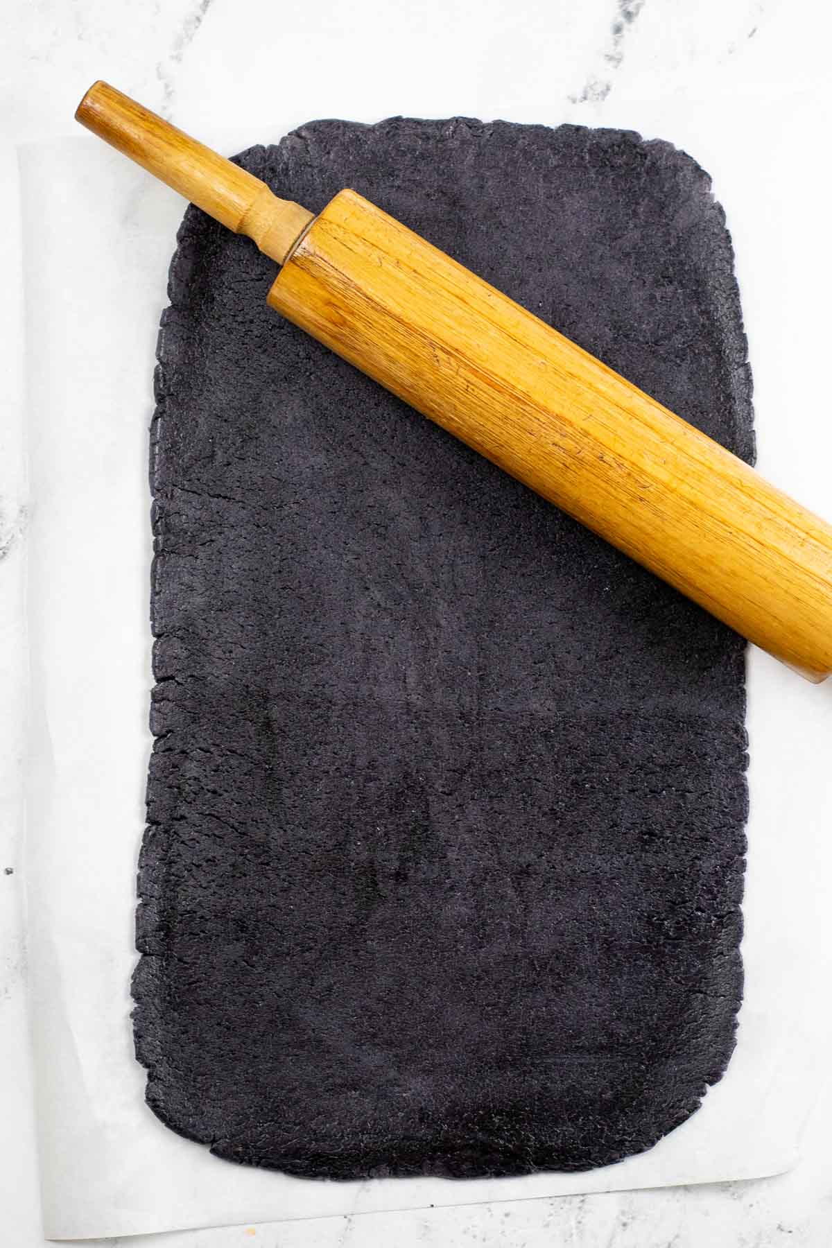 black cookie dough rolled out on a pice of parchment paper with a wooden rolling pin.