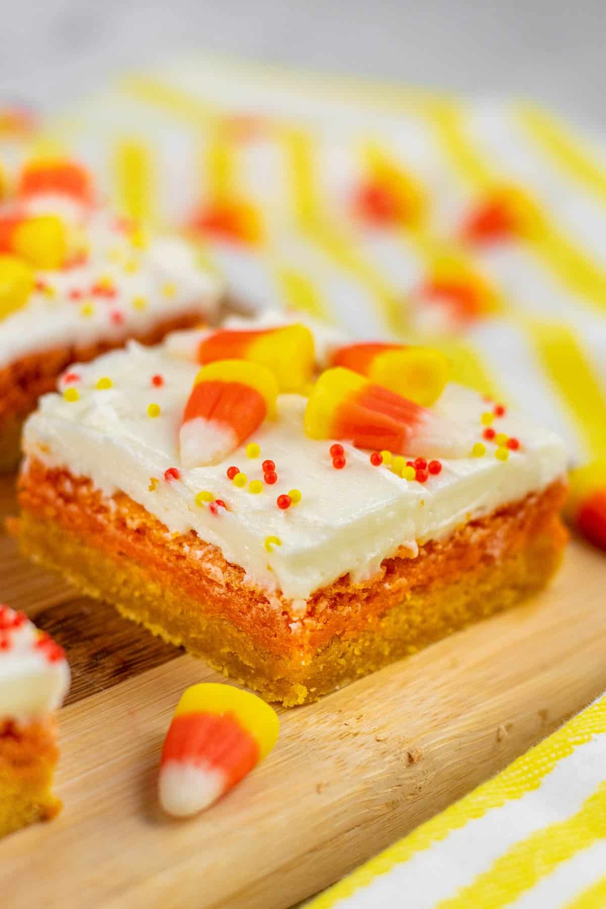 closeup of a orange and yellow layered sugar cookie bar topped with candy corn on a wood cutting board.