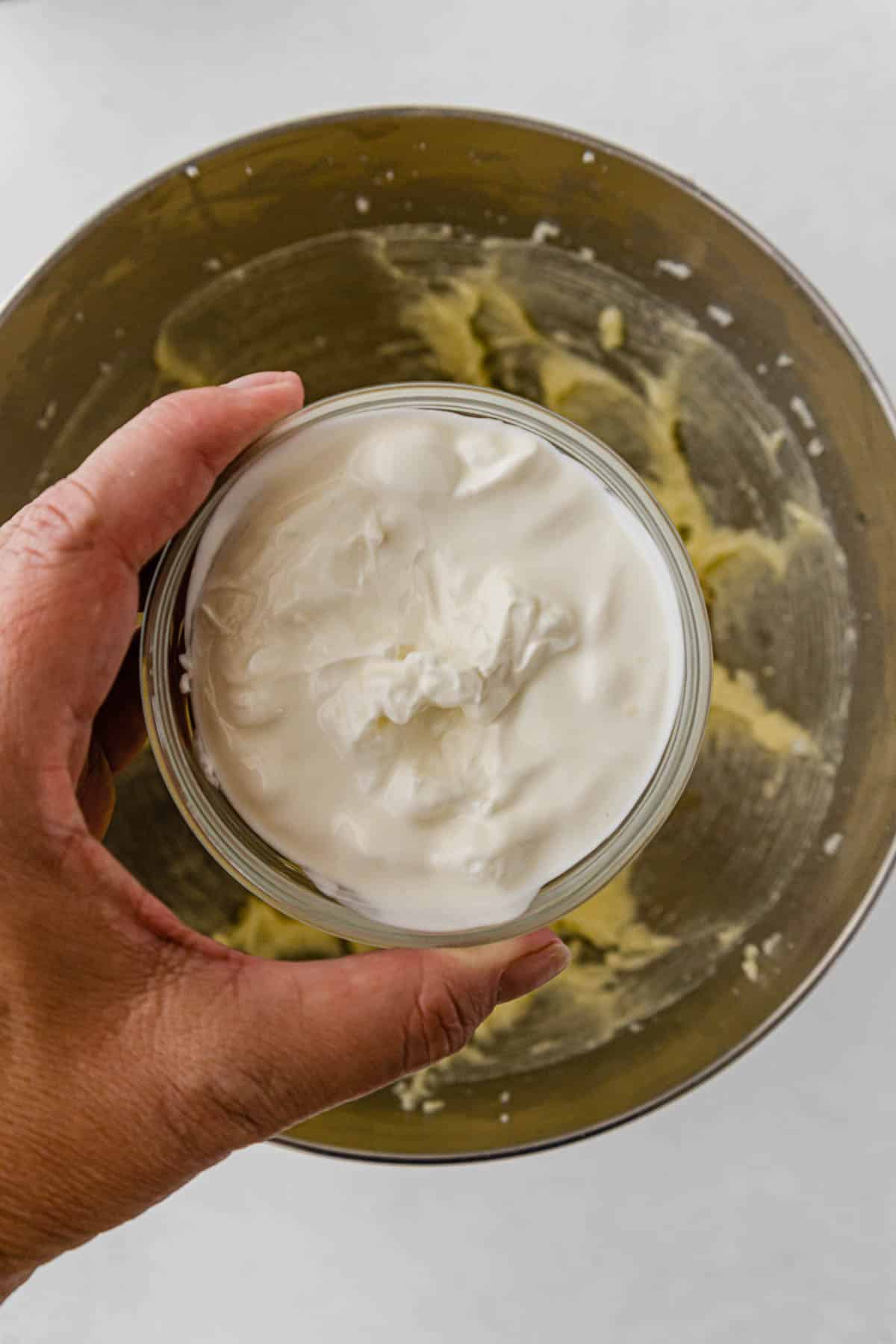 woman's hand holding a glass bowl of sour cream over a silver mixing bowl.