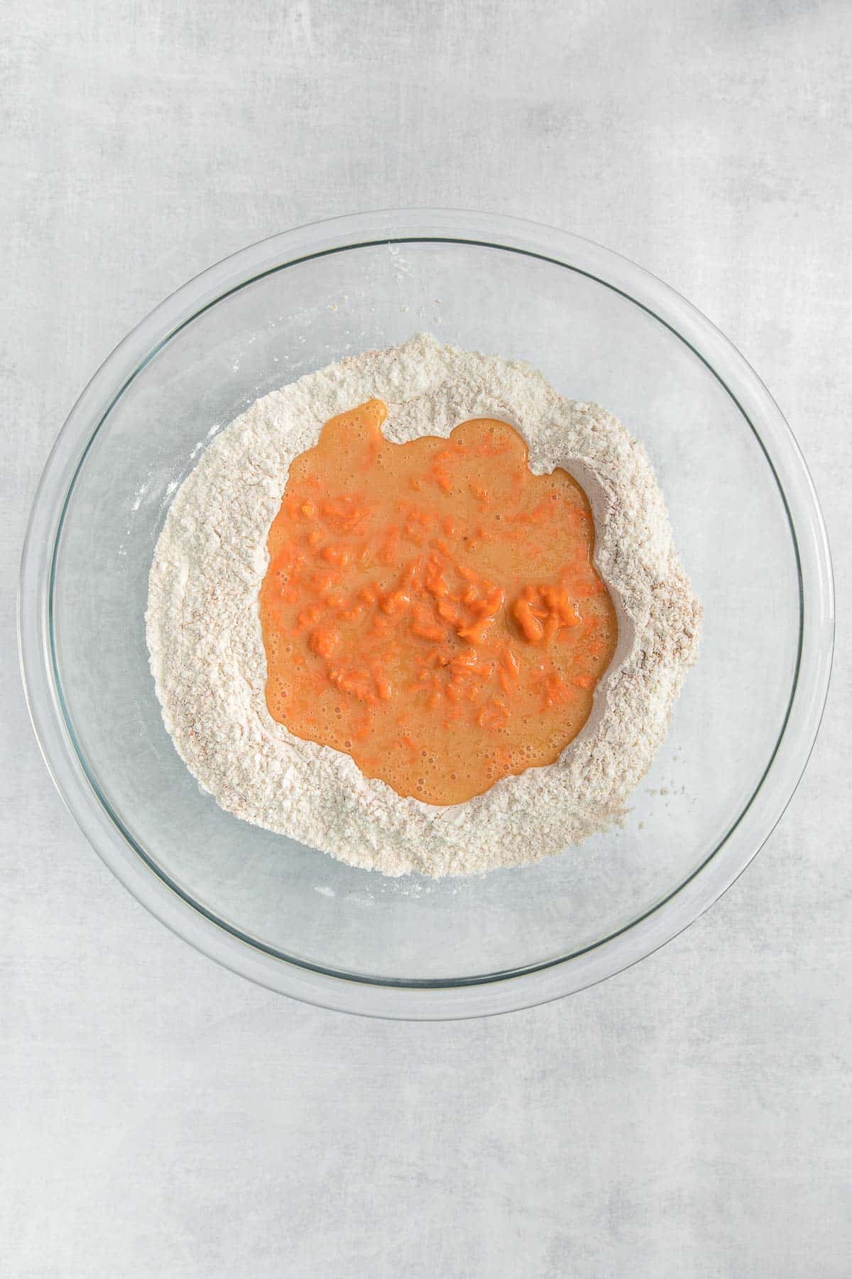 big glass bowl with flour topped with carrot and egg mixture.