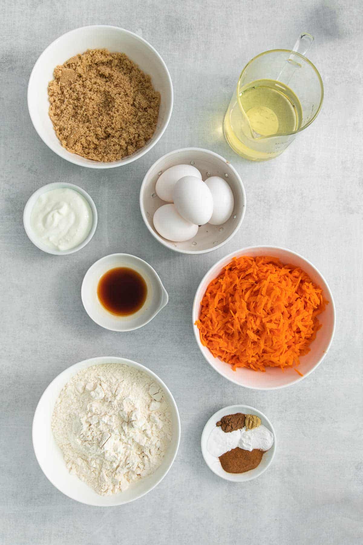several white bowls with ingredients for carrot cake - flour, oil, brown sugar, oil, eggs, grated carrots, spices,