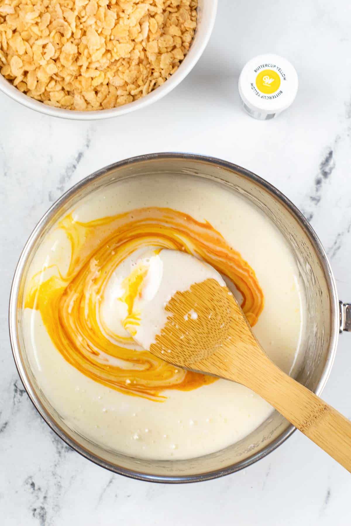 melted marshmallows in a silver pot with with yellow food coloring being stirred in with a wood spoon stirring.