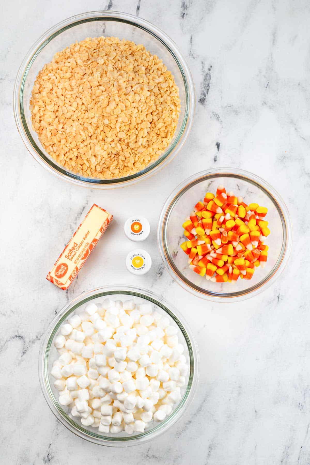 three glass bowls - one with mini marshmallows, one with candy corn and one with rice krispie cereal, a stick of butter