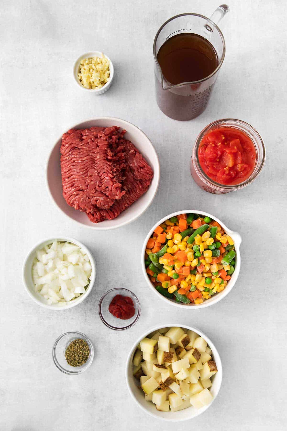 several white bowls with ingredients for hamburger soup - ground beef, diced potatoes, mixed vegetables, diced tomatoes, beef broth and seasonings.