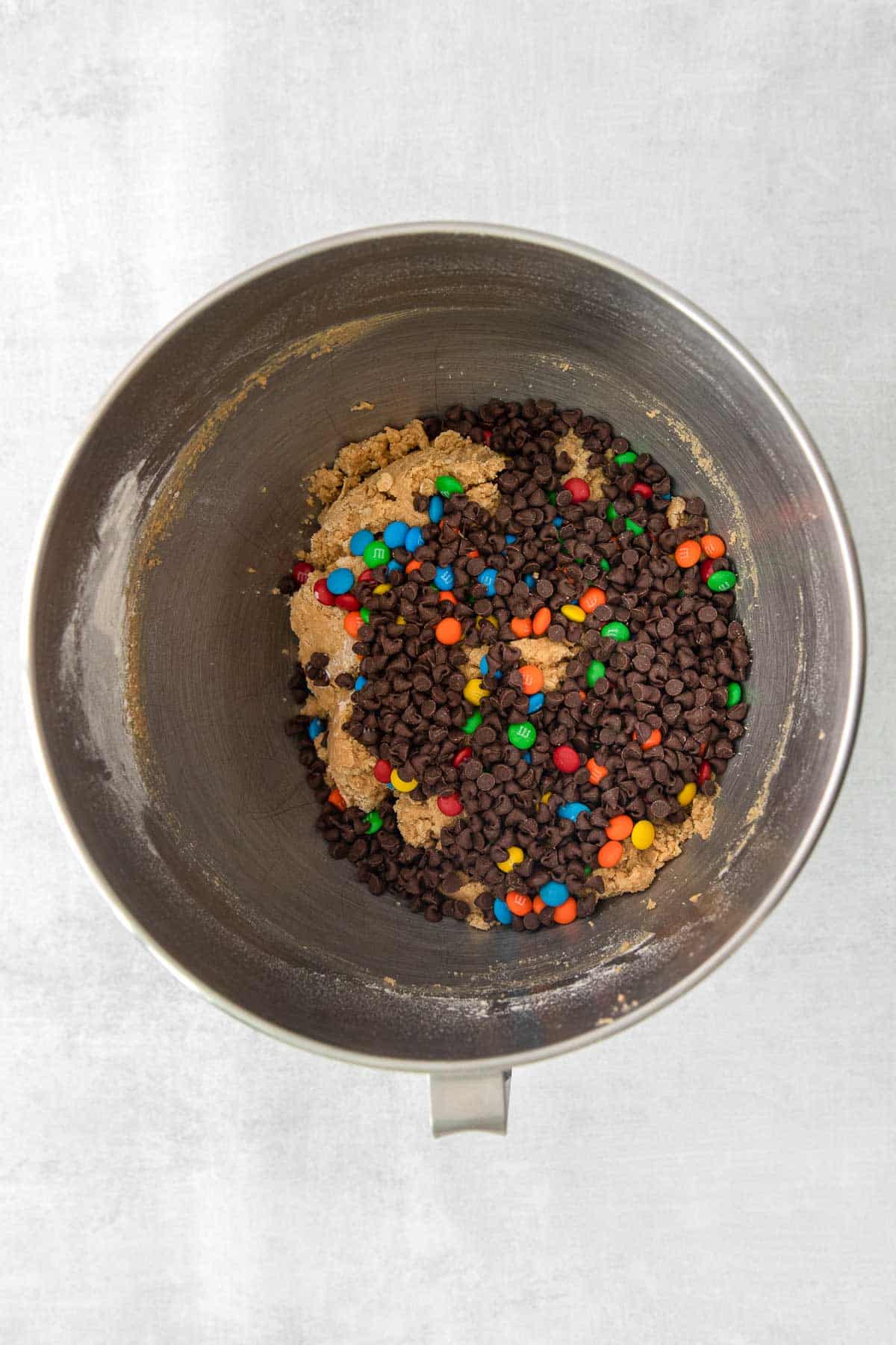 silver mixing bowl with chocolate chips and M&M candies on top of cookie dough.