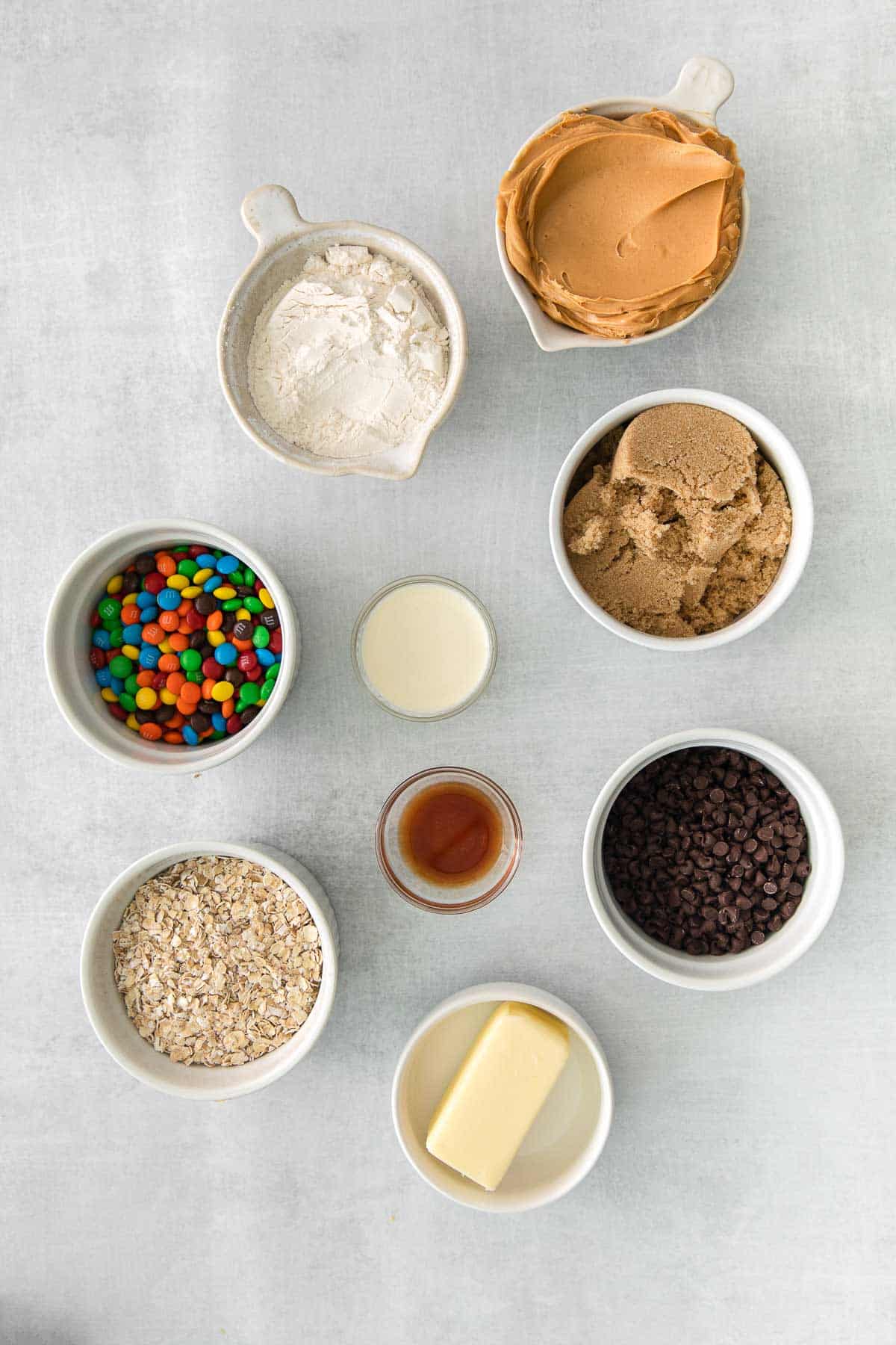 several white bowls with ingredients for monster cookie dough - oats, flour, butter, vanilla extract, peanut butter, chocolate chips, m&m's and milk
