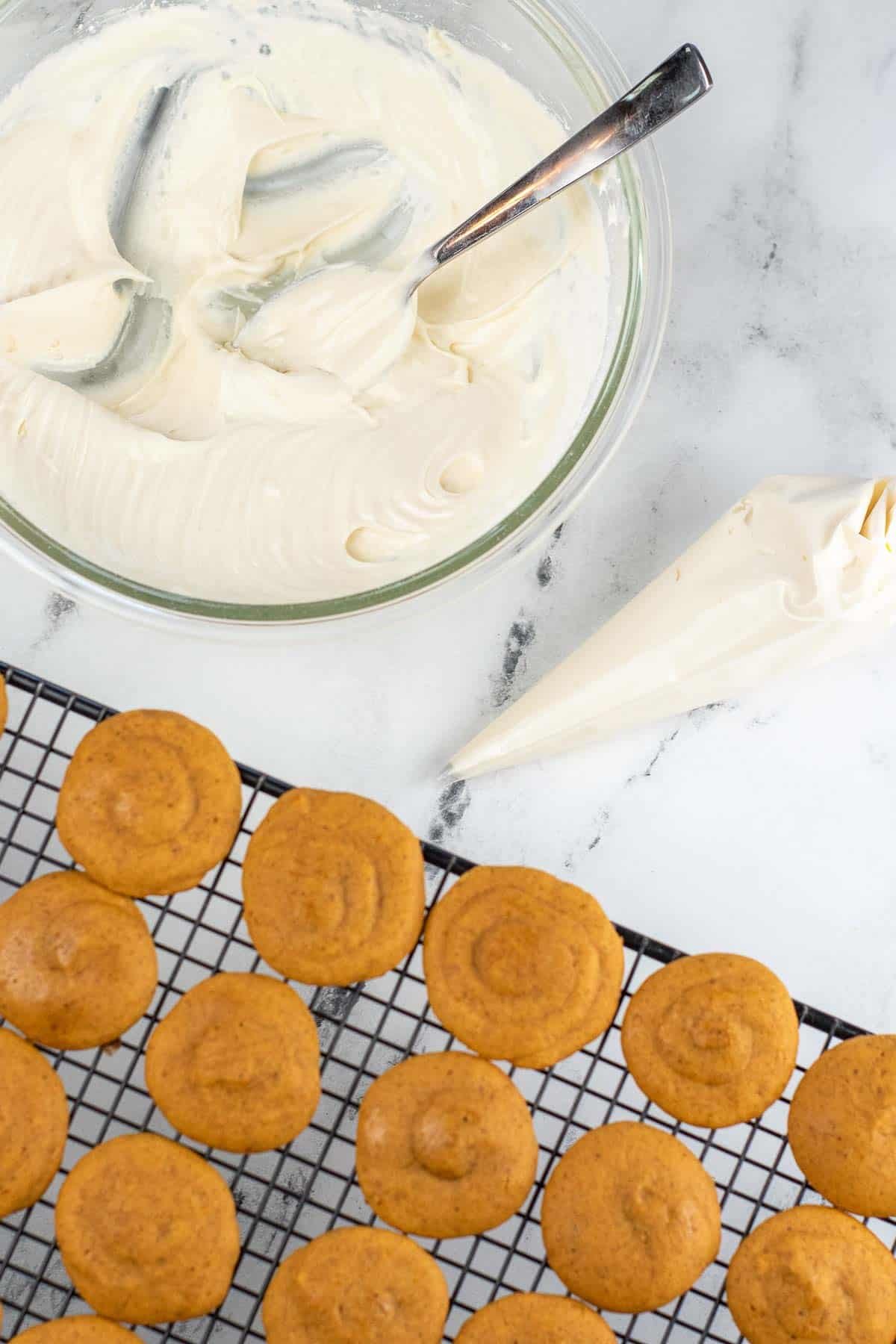 several pumpkin cookies on a wire cooling rack with a piping bag and bowl full of cream cheese frosting beside them.