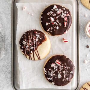 three cookies dipped in chocolate and topped with crushed peppermint candy on white parchment paper.