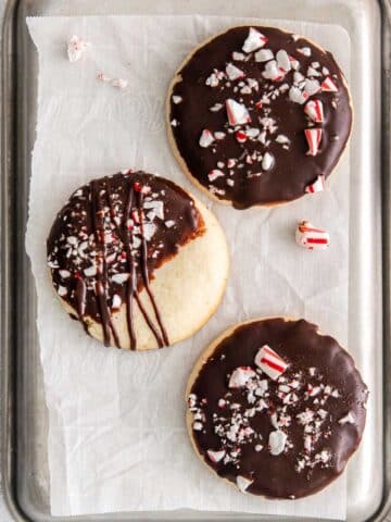 three cookies dipped in chocolate and topped with crushed peppermint candy on white parchment paper.