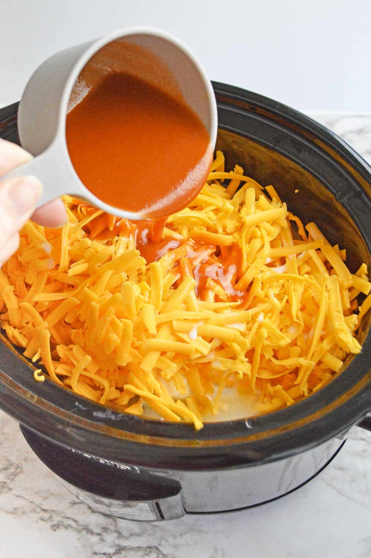 buffalo sauce being poured over shredded cheddar cheese in a crock pot.