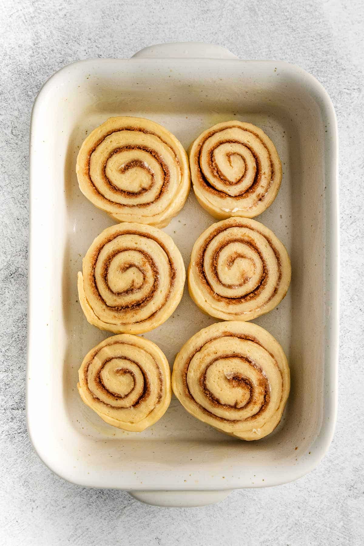 six uncooked cinnamon rolls in a white rectangle baking dish.