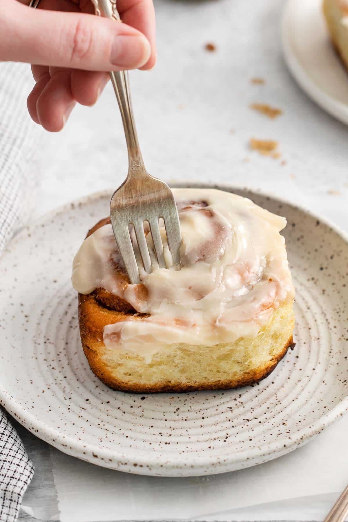 white plate with a cinnamon roll with white frosting with a fork sticking in the top.