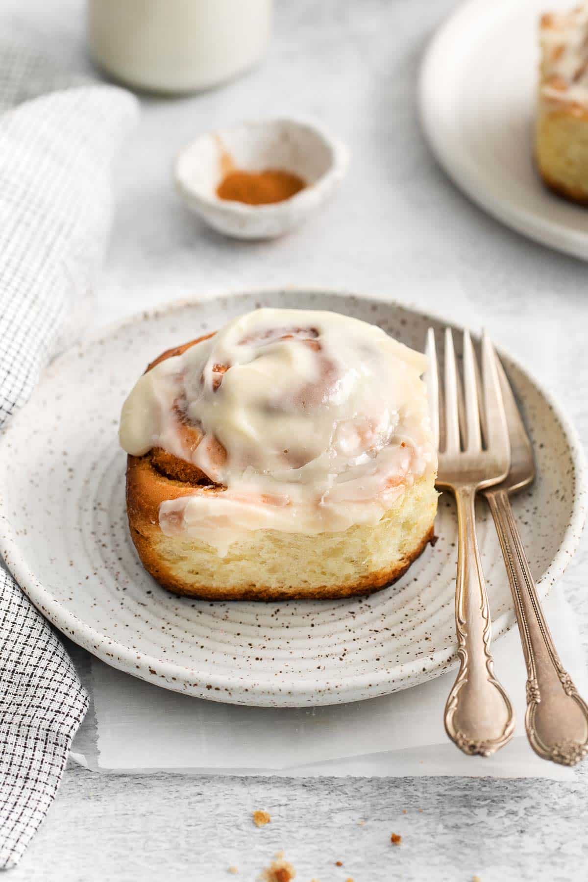 closeup of a cinnamon roll with thick icing on a white plate with two forks on the side of the plate.