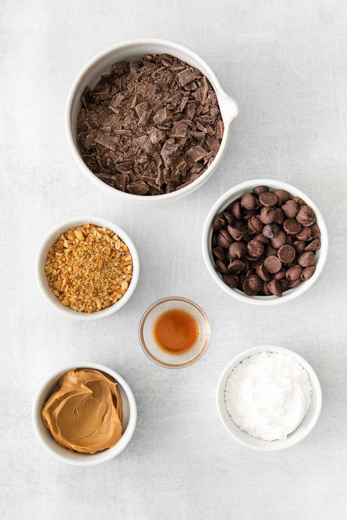 several white bowls with ingredients for homemade peanut butter cups creamy peanut butter, powdered sugar, vanilla extract, chocolate chips, chocolate bark and chopped peanuts.