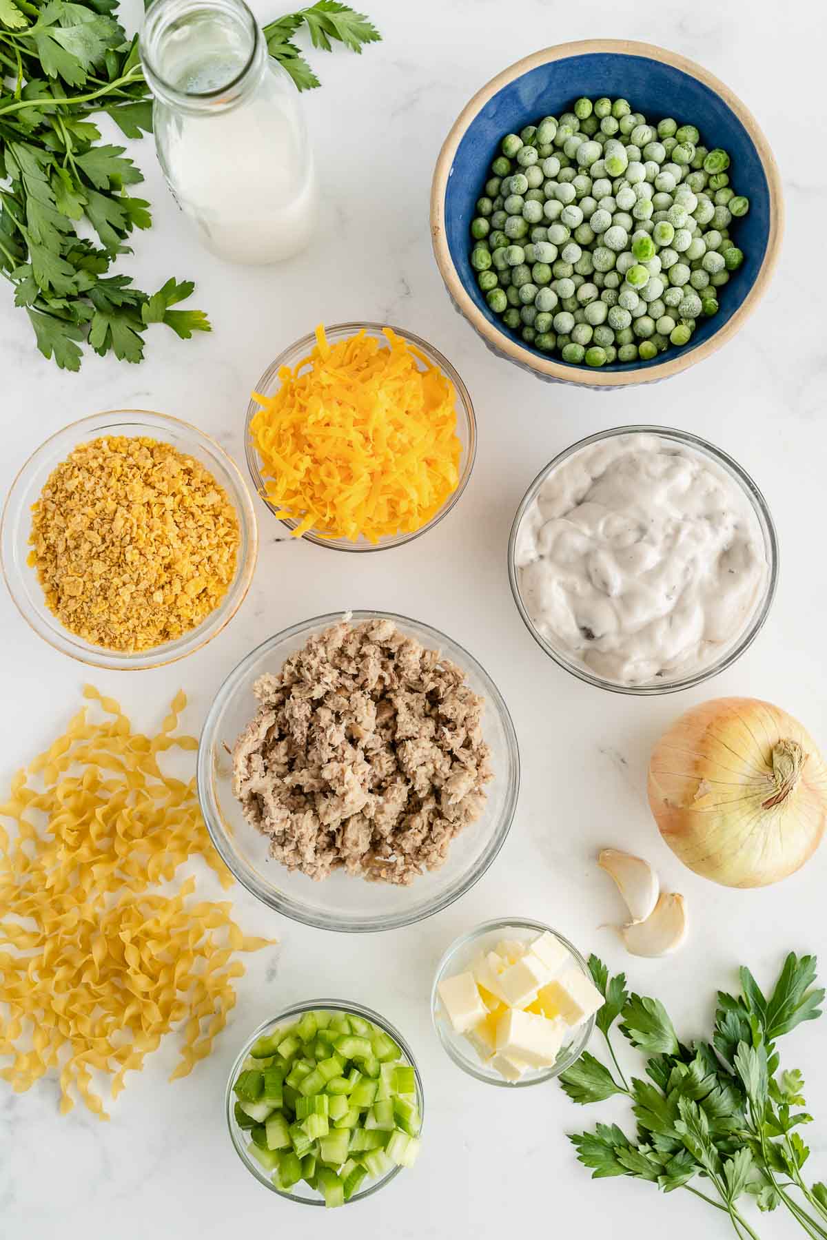 several bowls with ingredients for tuna casserole - canned tuna, onion, celery, egg noodles, butter, frozen peas, cream of mushroom soup, crushed cornflakes and shredded cheese.
