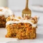 square slice of carrot cake with cream cheese frosting and chopped nuts with a fork in the top.