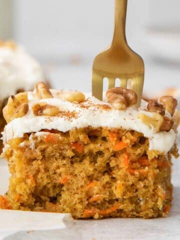 square slice of carrot cake with cream cheese frosting and chopped nuts with a fork in the top.