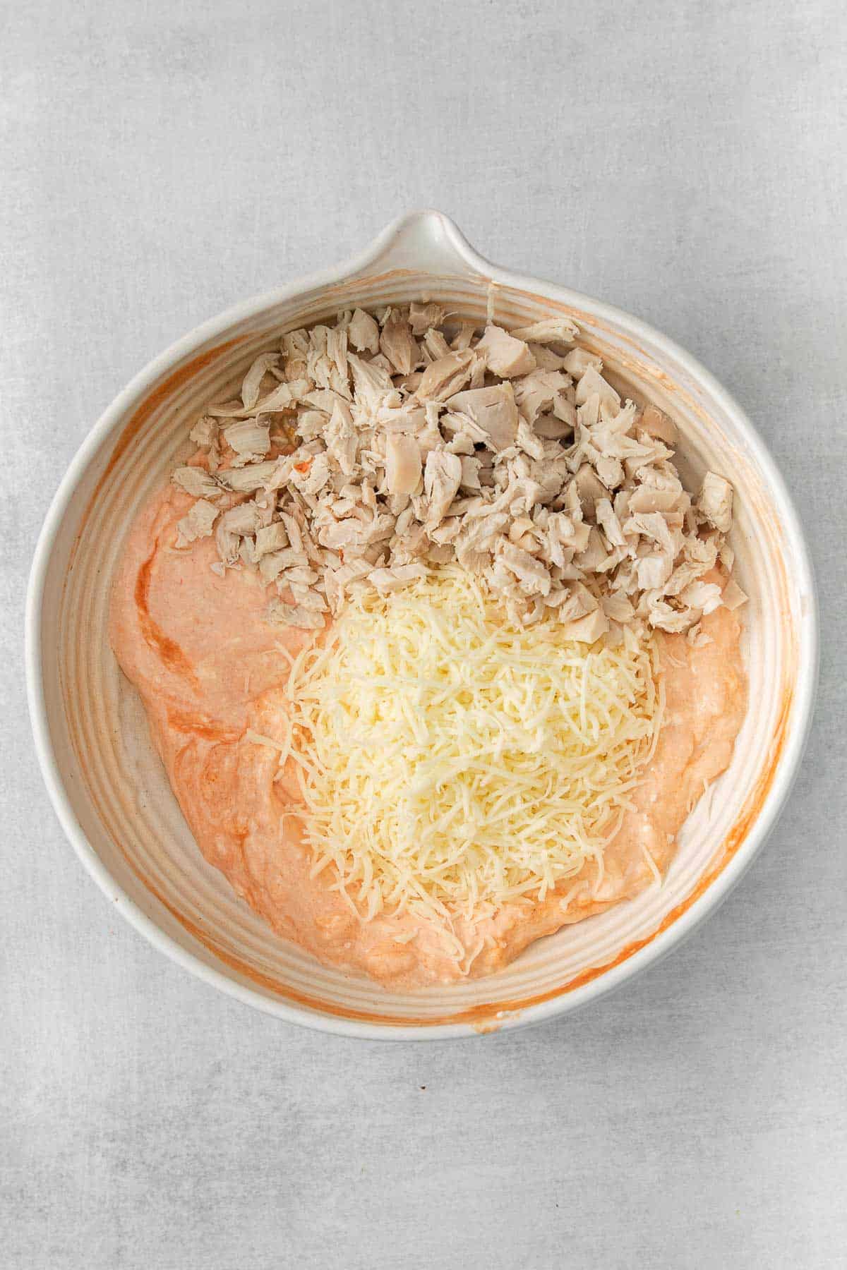 white mixing bowl with buffalo and cream cheese mixture with shredded chicken and shredded cheese.