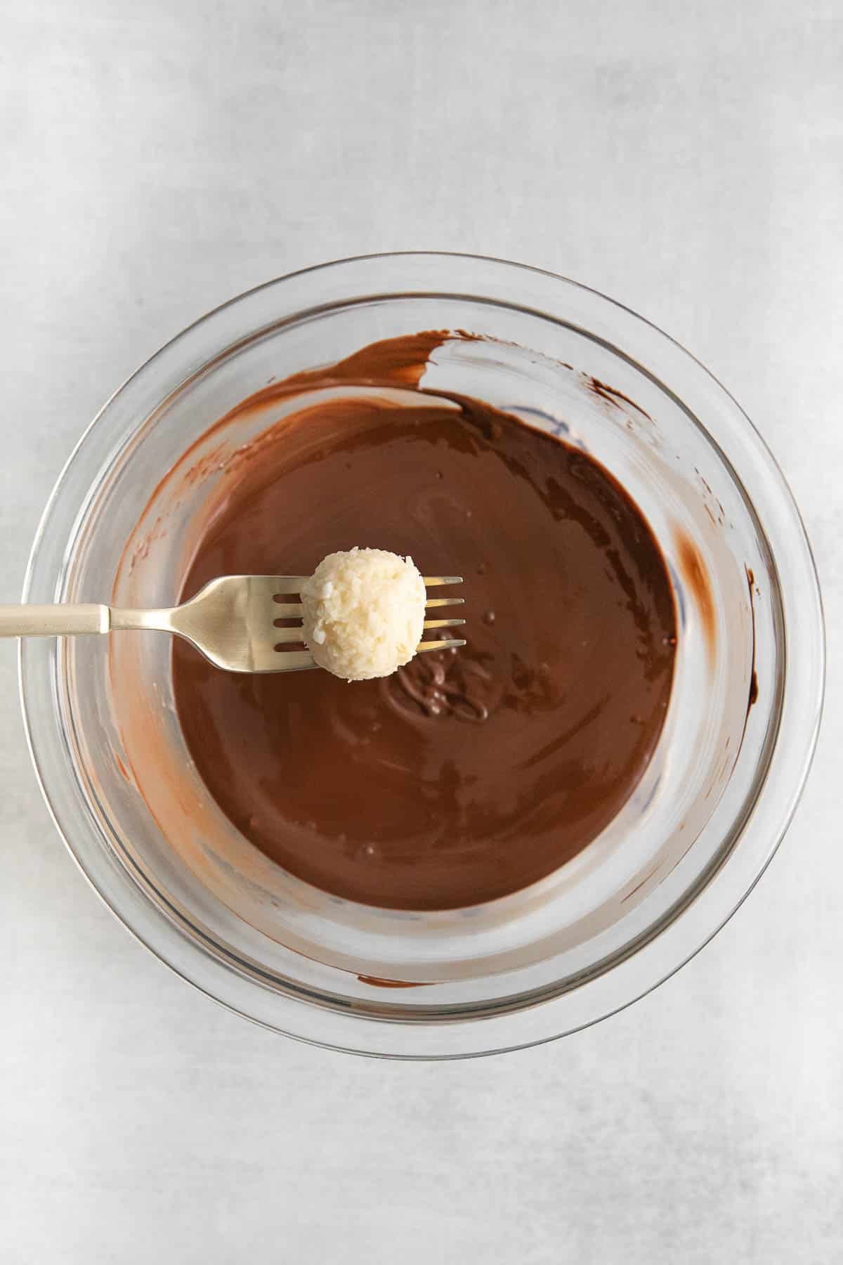 glass bowl of melted chocolate with a coconut ball on a fork being held over top of the bowl.