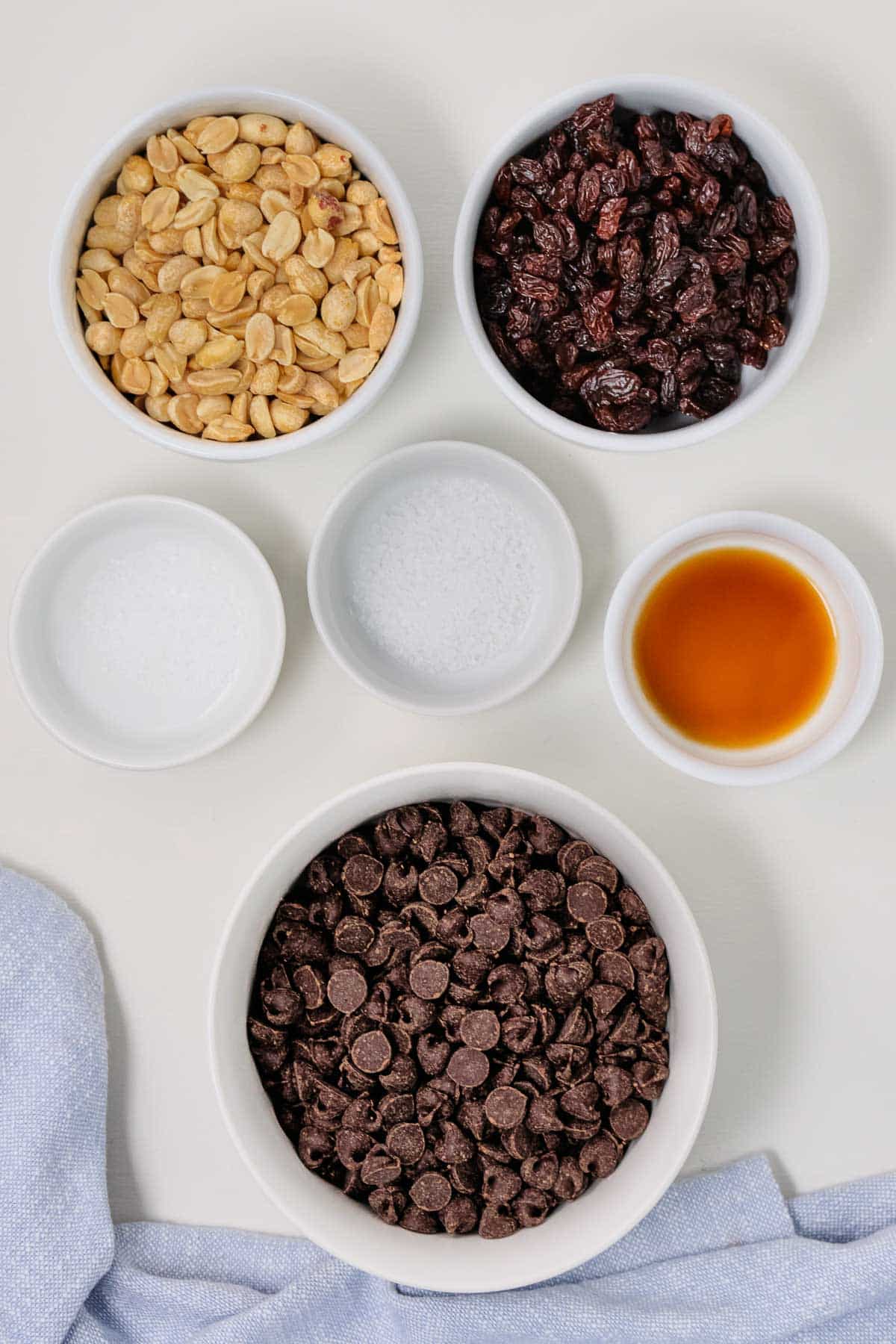 several white bowls with ingredients for chocolate covered peanuts - chocolate chips, peanuts, raisins, salt and vanilla extract.