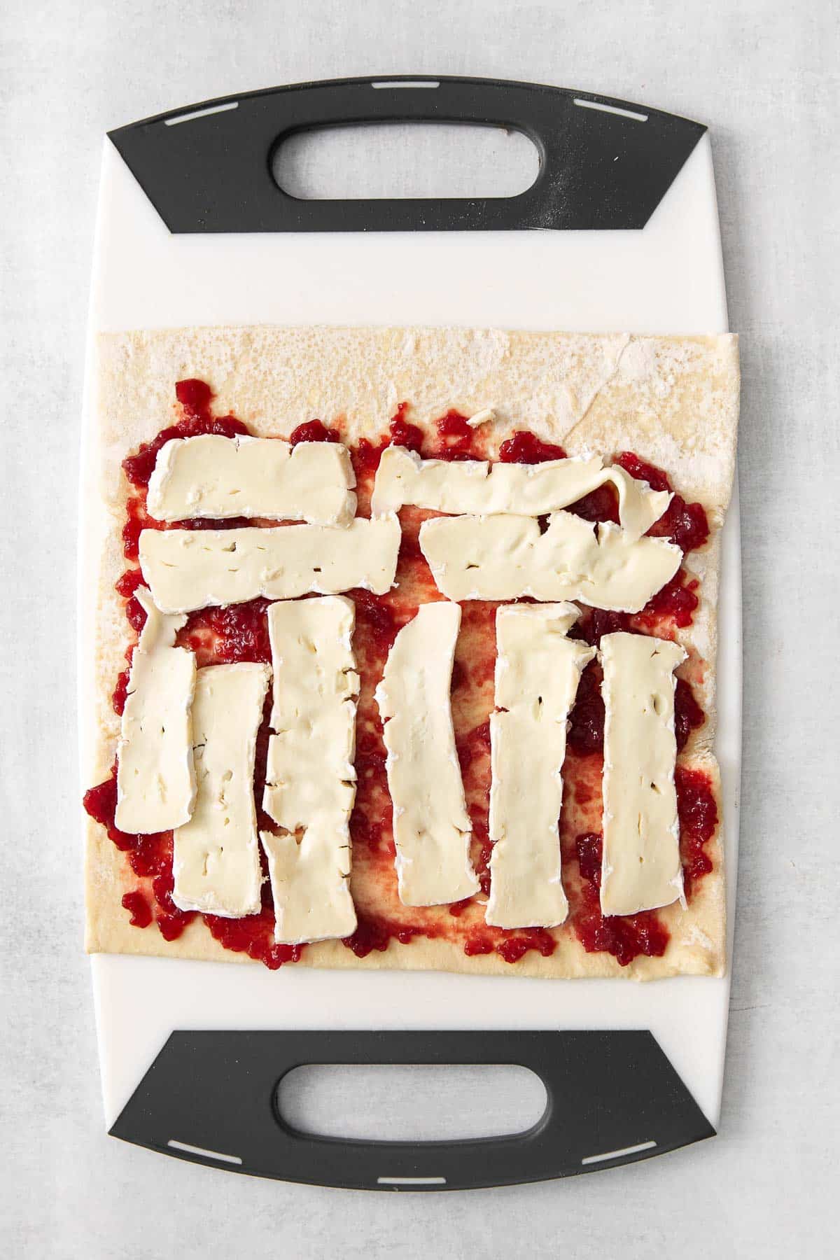 cutting board with puff pastry rolled out and cranberry sauce and sliced brie on top.