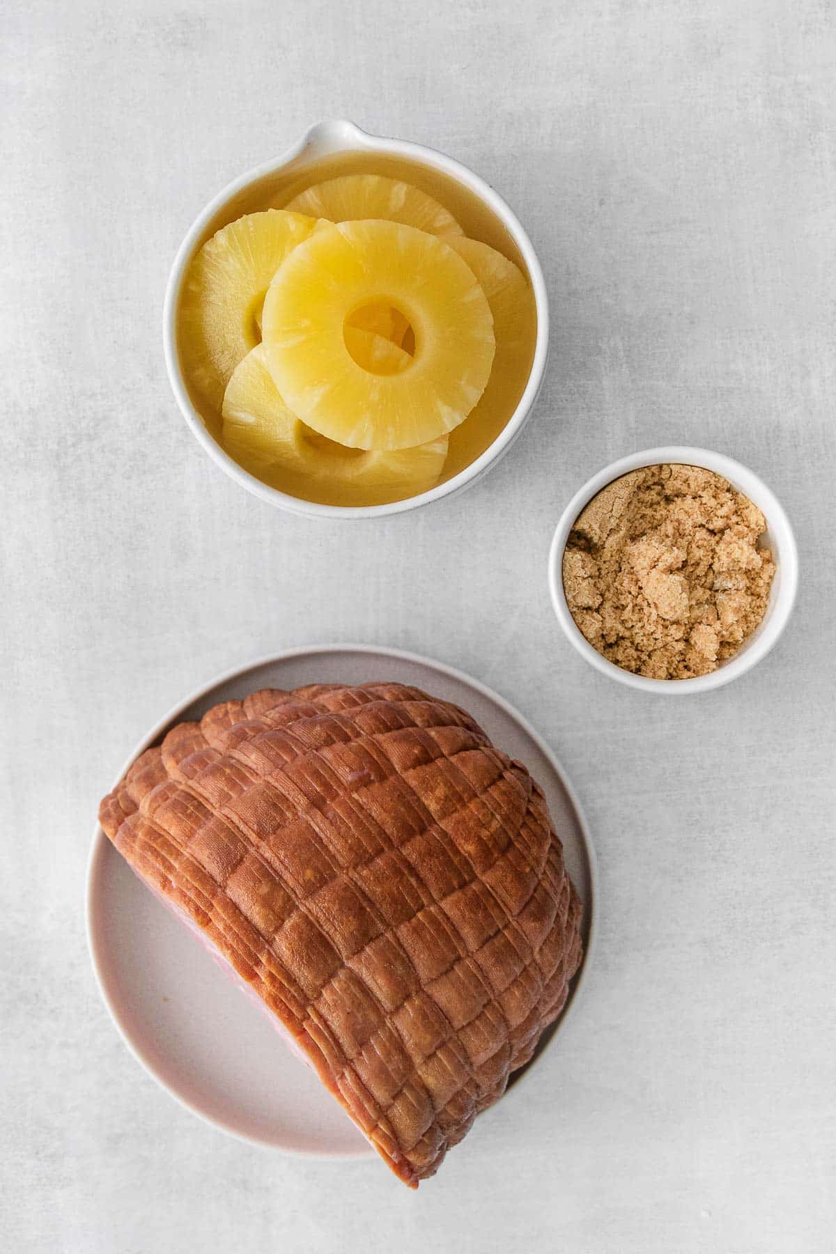 sliced ham on a plate and a bowl of pineapple rings and a small bowl of brown sugar.